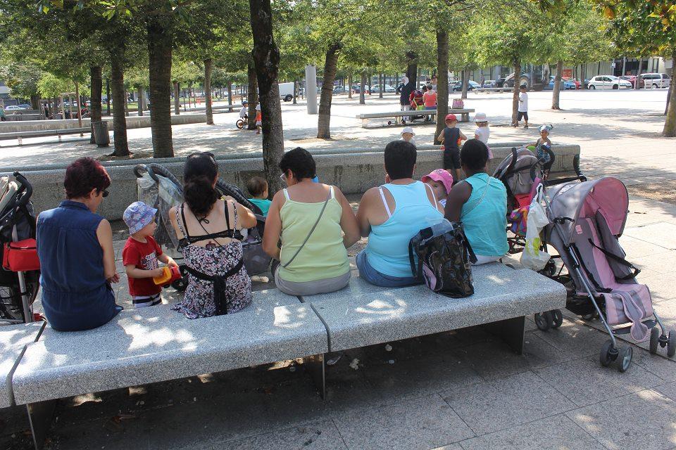 Plusieurs mères sont assises sur un banc Place des 5 fontaines. Elles sont entourées de poussettes et de leurs enfants dont elles s'occupent.