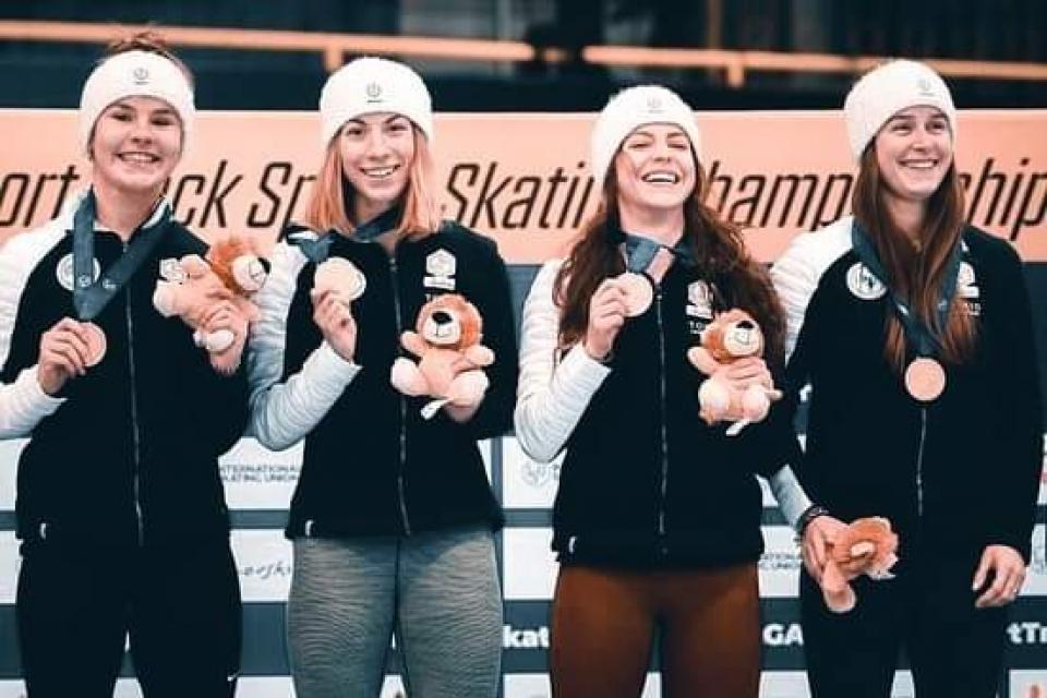 Photo prise de face, nous voyons, Aurélie Leveque, championne d'Europe de short track, avec ses 3 co-équipières. Elles sont toutes souriantes et tiennent leur médaille dans une main.