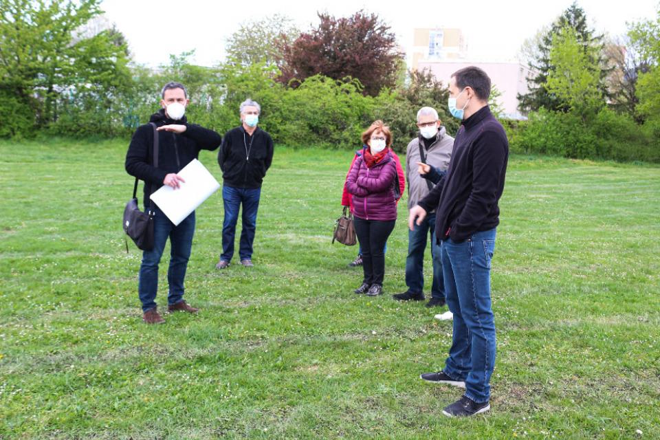 5 personnes masquées se tiennent dans un parc et discutent. Marco Rossi, l'architecte en charge du projet, explique les futures orientations aux habitant-es sur le terrain, grandeur nature.