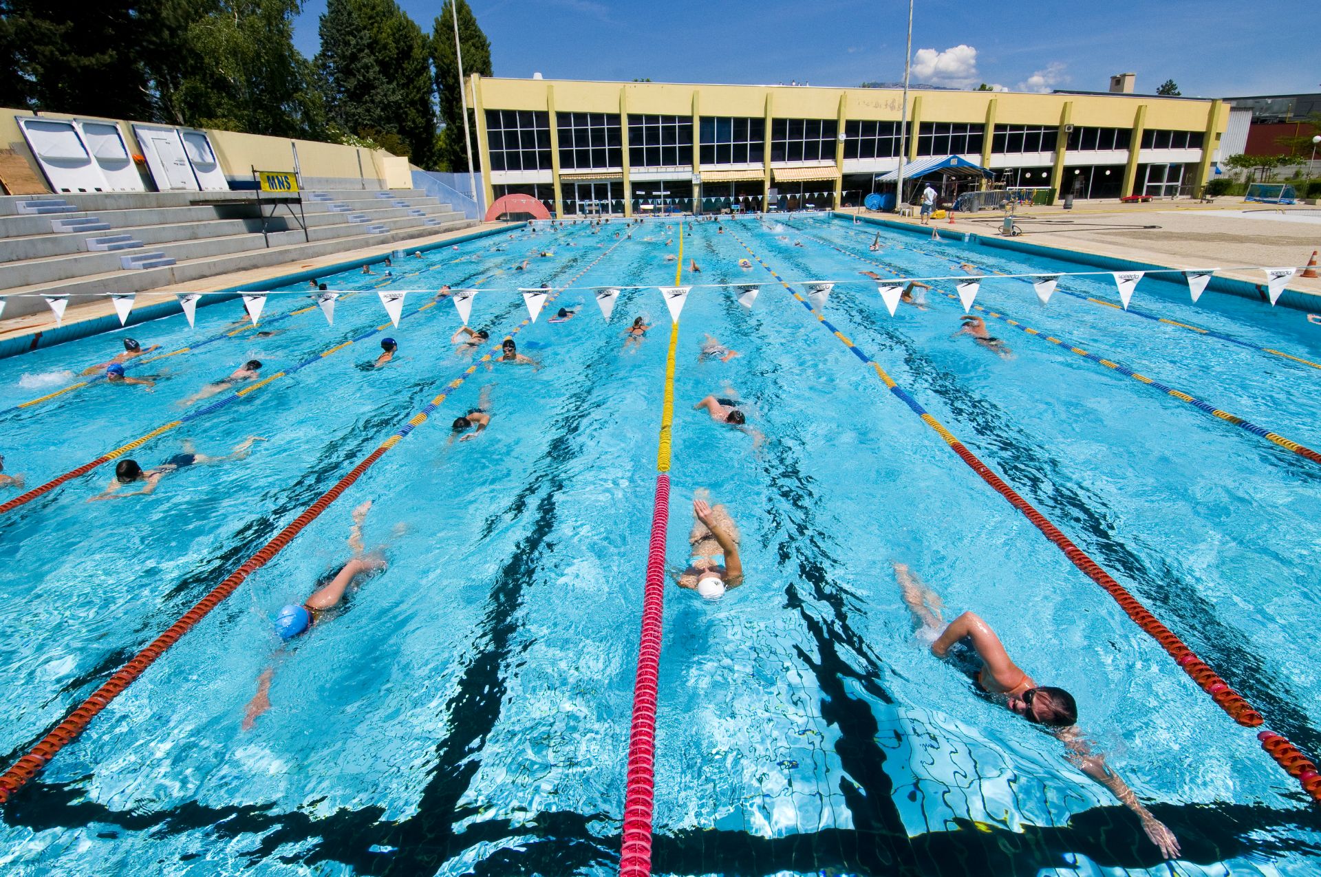 Bassin olympique extiéreur Vue sur le bassin extérieur du stade nautique. Plusieurs nageurs et nageuses occupent les lignes d'eau et font des allers-retours.