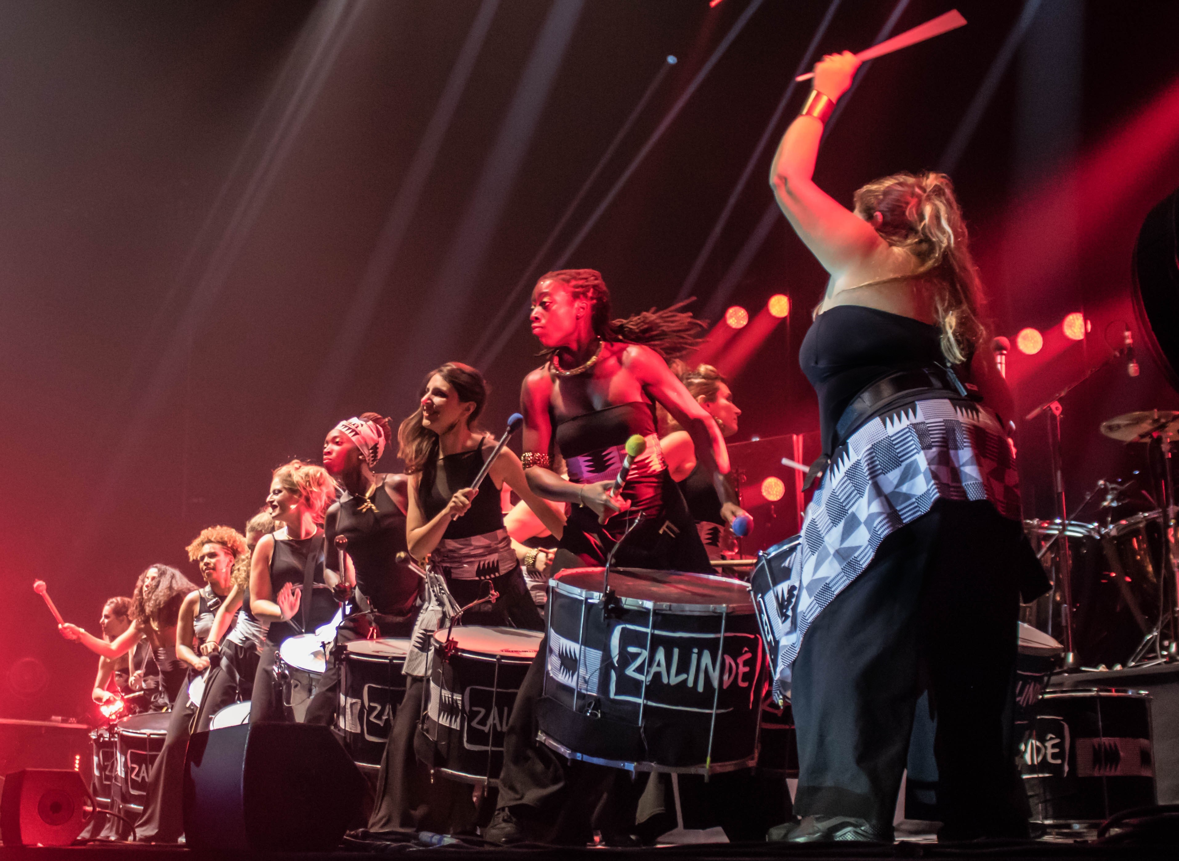 Vue sur la troupe de percussions. Les musiciennnes sont toutes habillées de noire, avec une ceinture de couleur. Elles ont les tambours devant elles. Il est écrit "zalindé" sur chaque tambours.