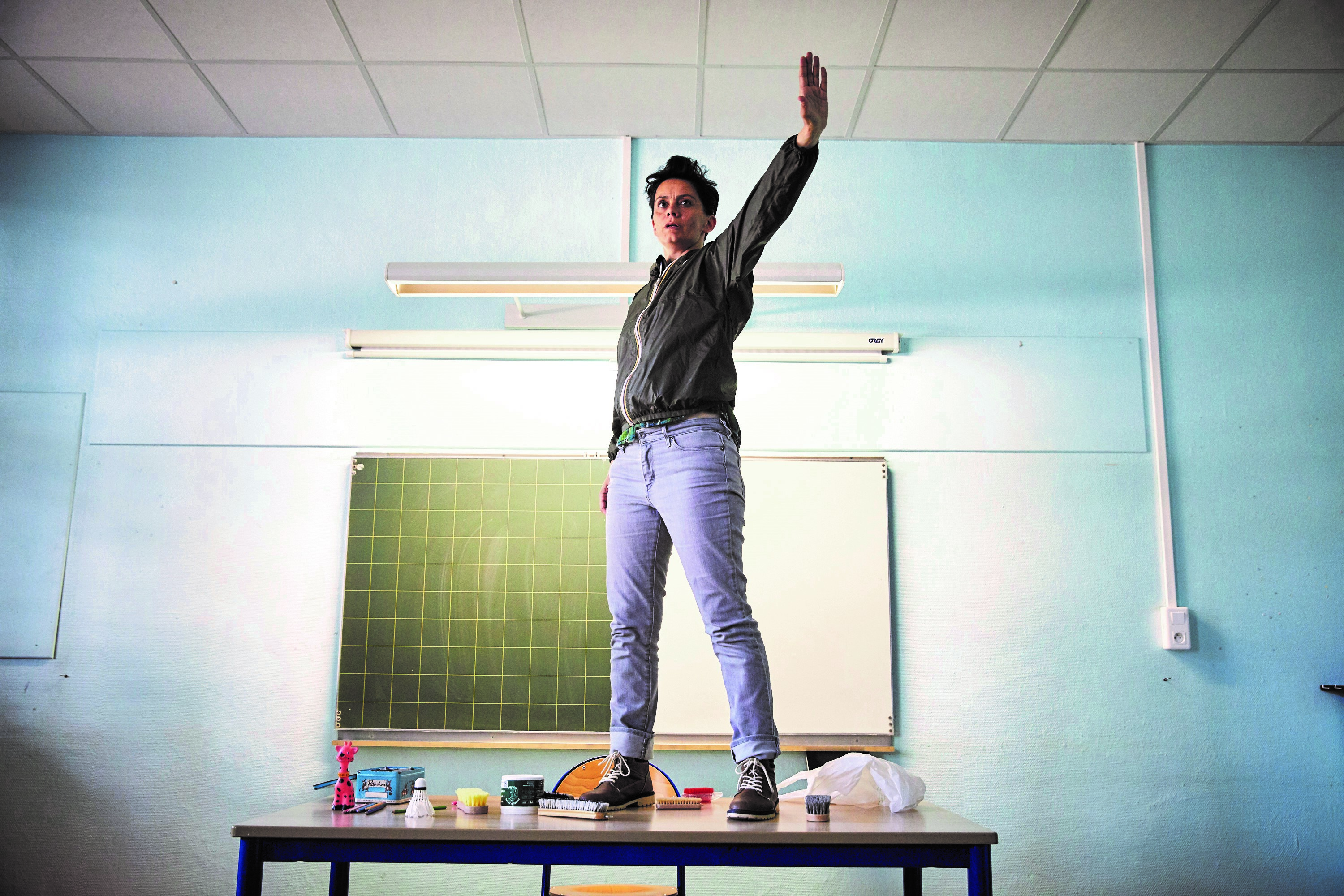 Elle pas princesse, lui pas héros ; Maia Le Fourn (c) Christophe Raynaud de Lage La photo est prise dans une salle de classe. Un acteur se tient debout sur le bureau, le bras gauche levé d'une manière revendicatrice. On aperçoit le tableau derrière lui.