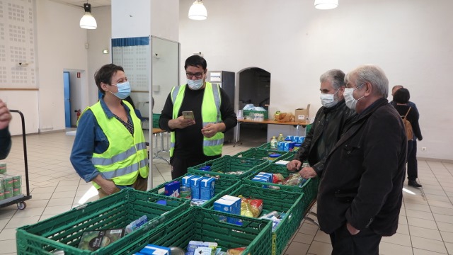 Distribution alimentaire de l'association Nouveaux horizons En cette période de confinement, le maire, masqué, visite le lieu d'un distribution alimentaire. Plusieurs cagettes de produits alimentaires sont alignés sur une table. De l'autre côté de la table, des bénévoles, masqués eux aussi, organisent la distribution.