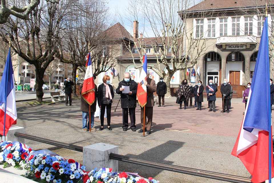 Vue sur la Commémoration du 19 mars 1962 - Cessez-le-feu en Algérie sur la place de la Libération.  Jean-Pierre Falque et Georges Chaix, masquéz, réalisent un appel aux morts afin que la mémoire des victimes de la guerre d'Algérie, des combats en Tunisie et au Maroc perdure. Ils sont entourés par 2 portes drapeaux et se tiennent face au monument aux morts, au pied duquel sont déposées des gerbes de fleurs. En arrière plan nous voyons les élu-es de la Ville qui assistent à la lecture.