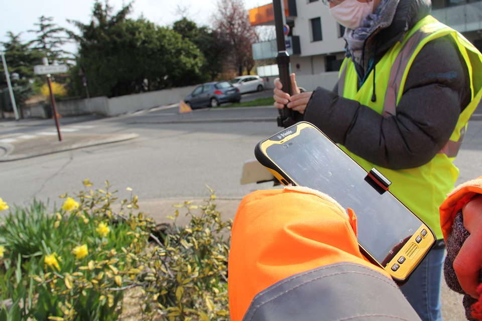 Vue sur deux agentes de la Ville qui effectuent un relevé précis des modifications apportées à la voirie, aux espaces verts, au petit mobilier, marquage au sol, panneaux de signalisation. Elles sont équipées d'un  GPS situé au sommet d'une perche de géomètre et d'une tablette qui enregistre les informations recueillies.