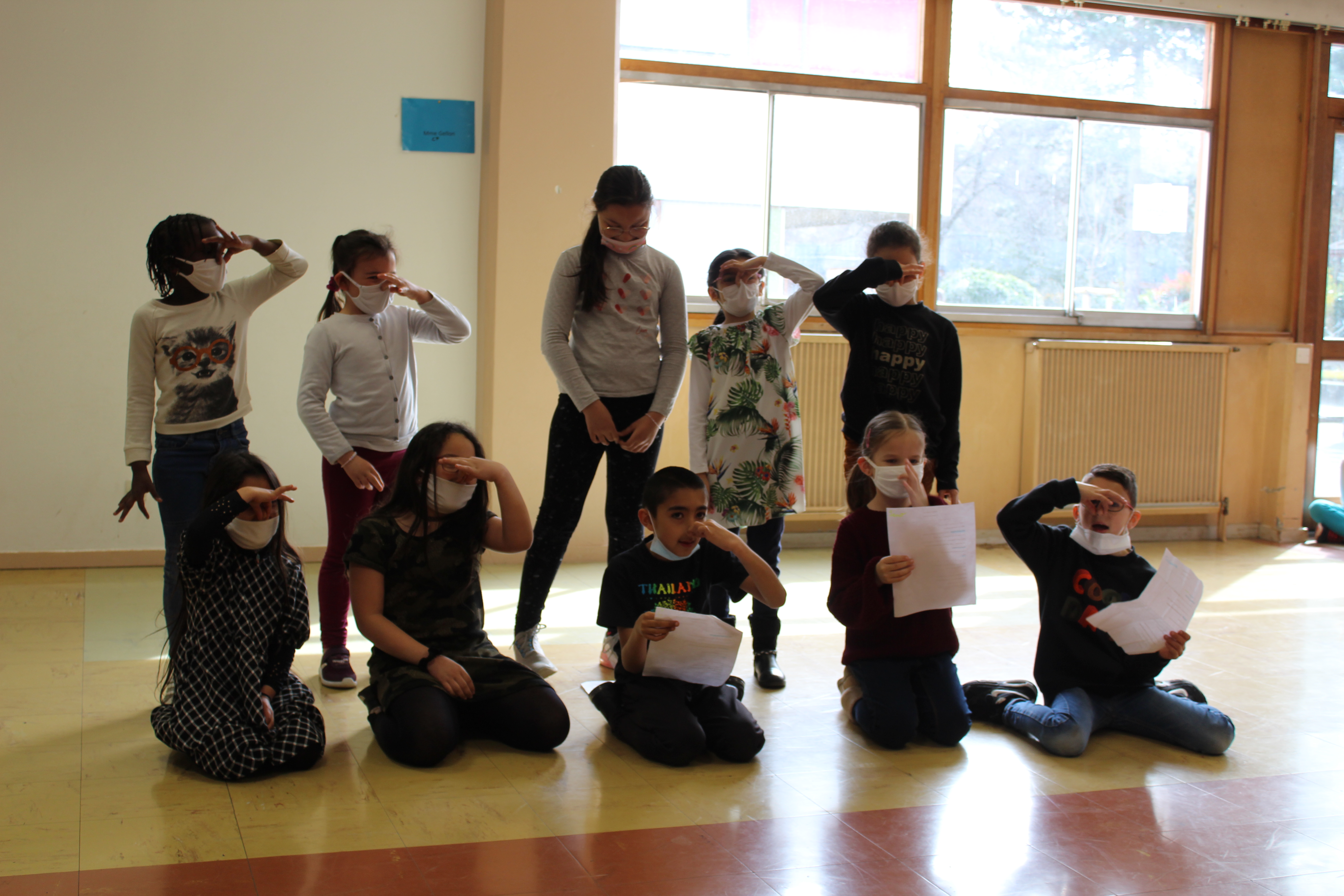 10 enfants font face à la photo dans le cadre d'un atelier théâtral. Ils sont répartis sur 2 lignes, 5 devant, 5 derrière. La ligne arrière est debout, celle devant est assise. Tous les enfants se bouchent le nez dans un geste théâtral.