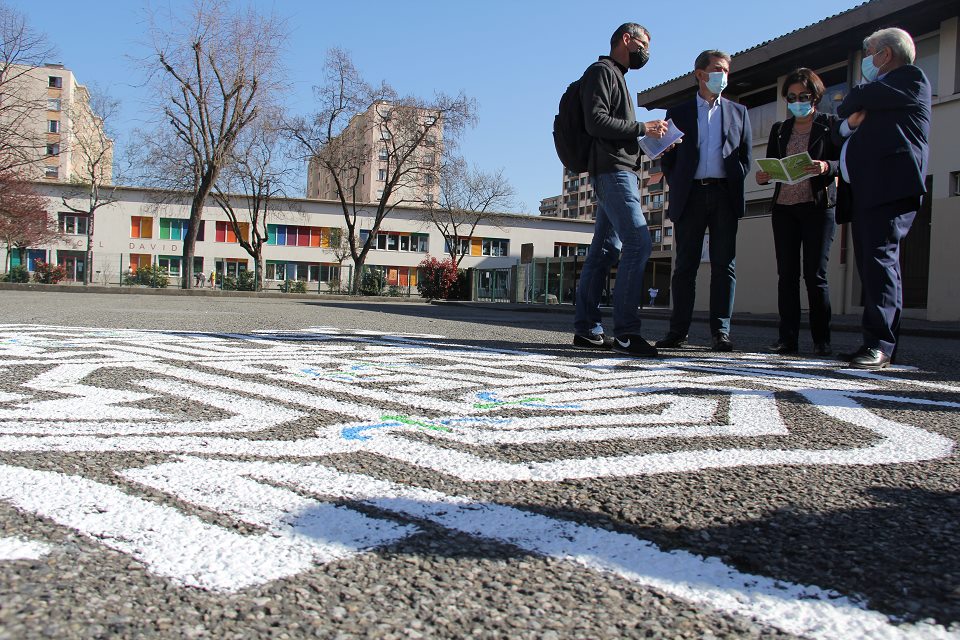 Photo prise en contre plongée. Vue sur le parcours d'énigmes dessiné sur le sol devant le gymnase, qui permettra aux enfants et aux parents de découvrir, en 9 étapes, les grands axes du projet. En arrière plan nous voyons les élu-es en discussion autour du projet.