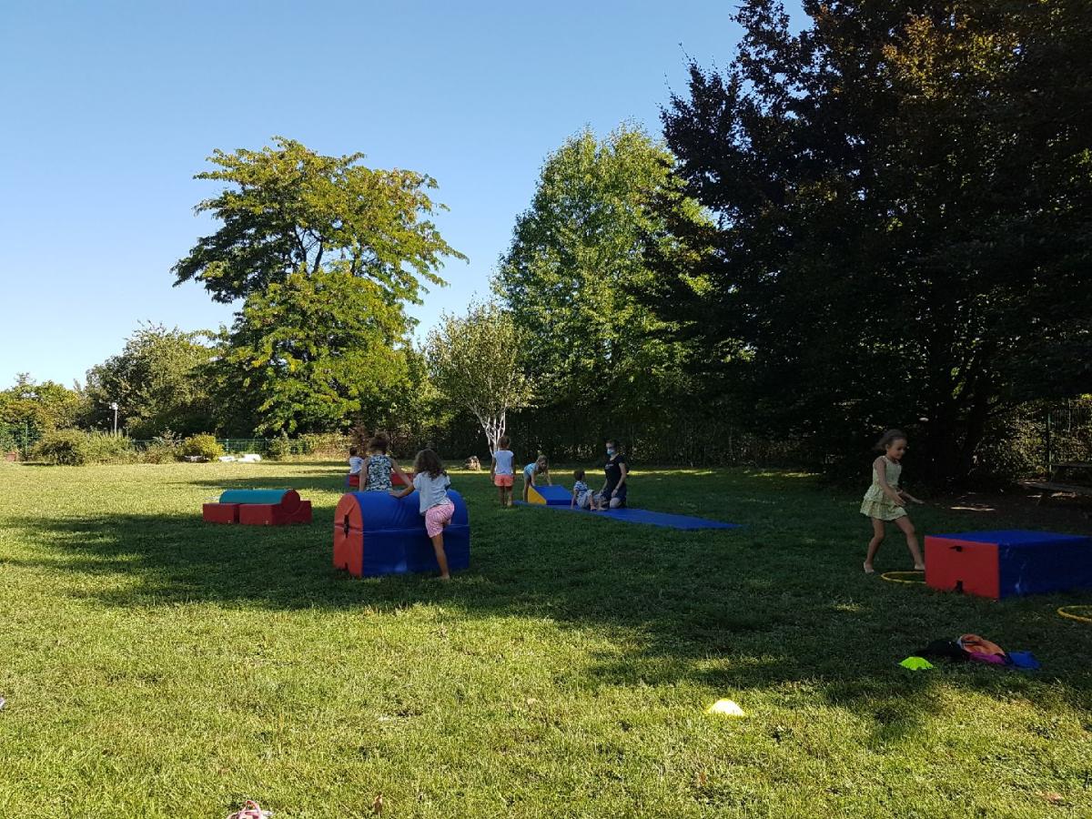 Des ateliers motricité sont organisés dans un parc de la Ville. Nous y voyons une accompagnatrice masquée. Les enfants marchent dans des cerceaux, enjambent des obstacles volumineux en mousse, etc.