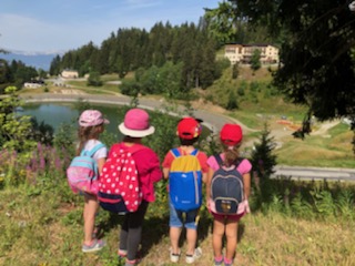 Sortie nature à évade Photo de 4 enfants prise de dos. Chaque enfant porte sa casquette et son sac à dos. Ils font face à un lac de montagne en contre bas de la photo.