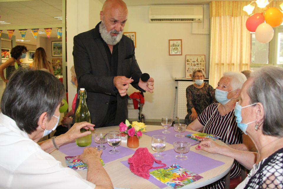 Photo prise à la fête de l'été de la Résidence autonomie Maurice-Thorez. Nous voyons les résidant-es seniors assis autour de tables rondes. des ballons colorés sont suspendus au plafond. Les résidant-es assistent à un spectacle de magique.