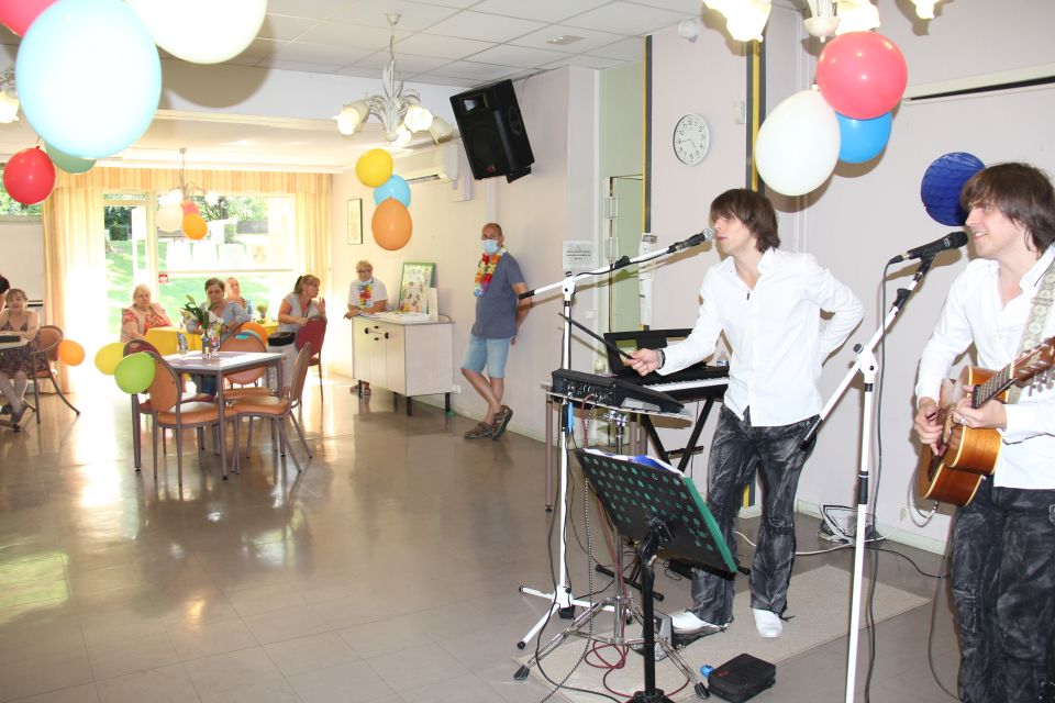 Photo prise à la fête de l'été de la Résidence autonomie Maurice-Thorez. Nous voyons les résidant-es seniors assis autour de tables rondes. des ballons colorés sont suspendus au plafond. Les résidant-es assistent à un spectacle de musique.