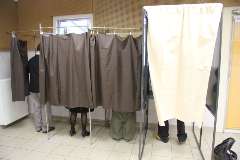 Photo prise lors d'une élection. Nous voyons les pieds des électeurs, sous le rideau de 4 isoloirs.
