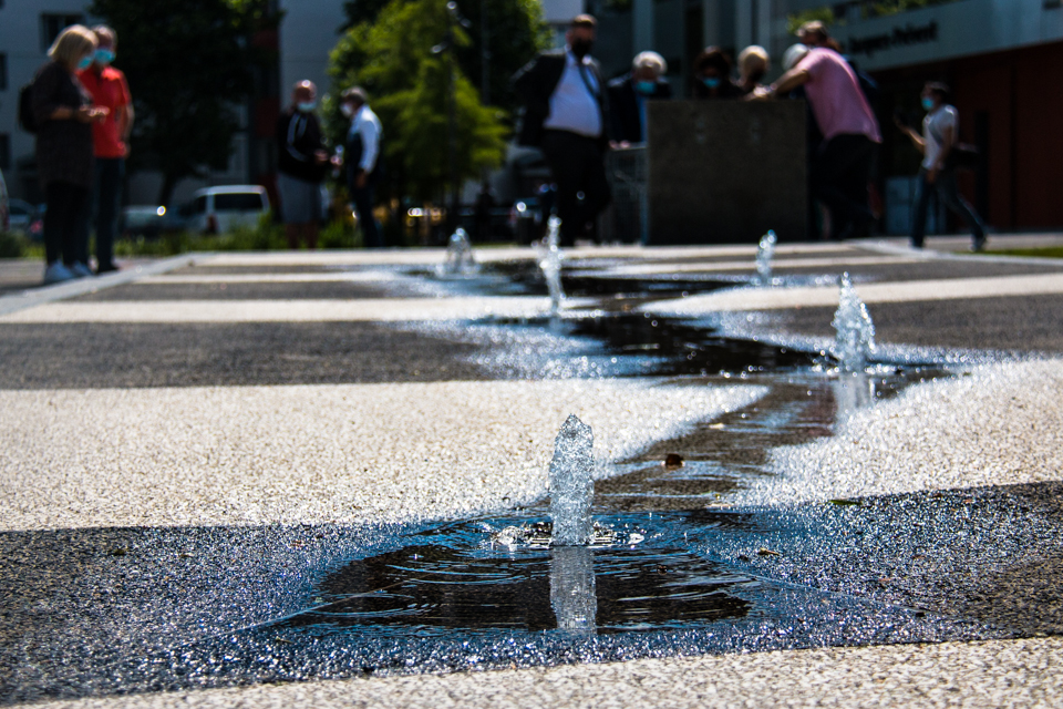 Photo prise dans le cadre de la remise en eau des fontaines 2021. Nous voyons les six jets de la fontaine du village 2 au premier plan. En arrière plan nous voyons des élu-es et des habitant-es.