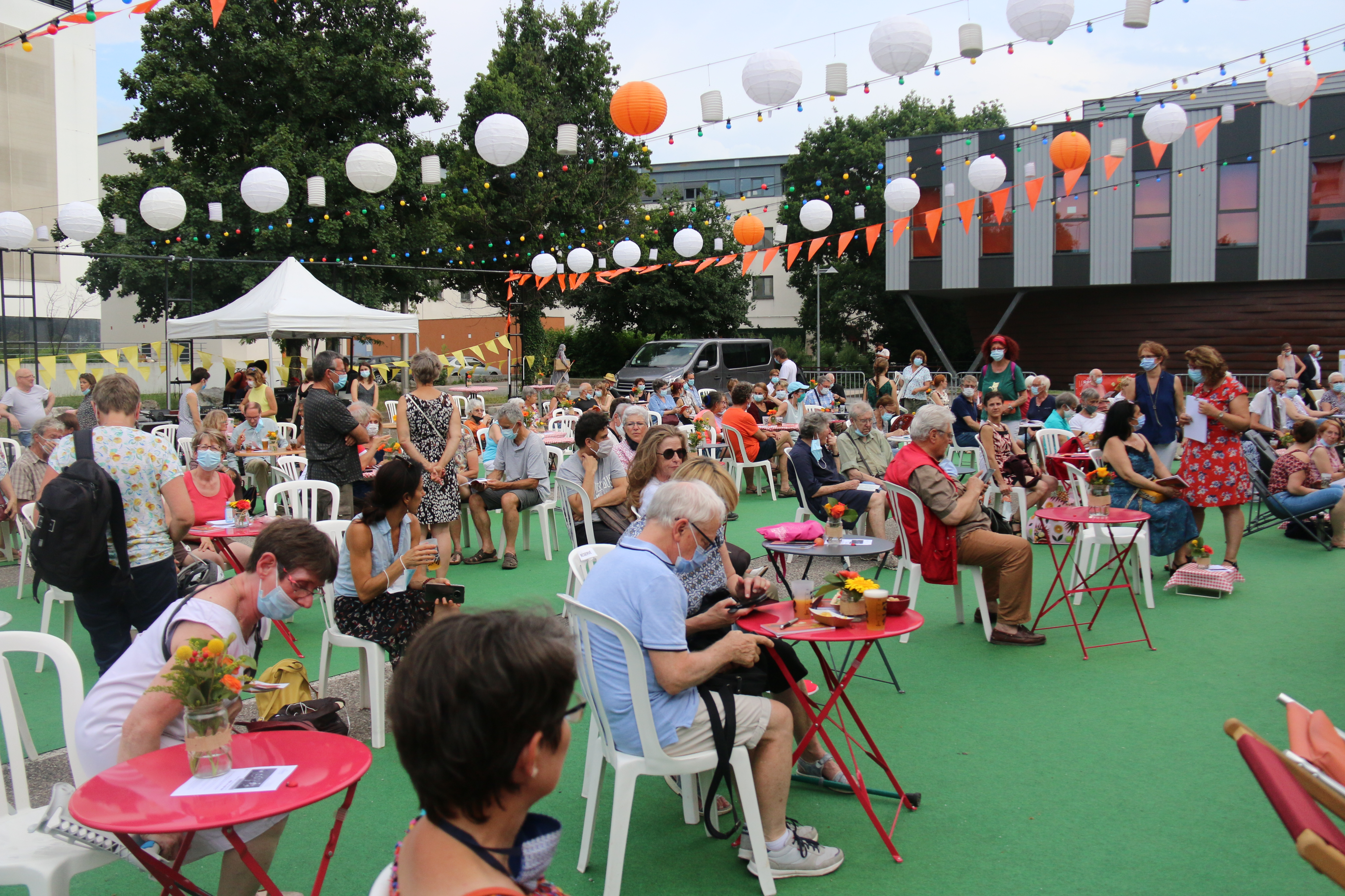 Photo de la présentation de saison la Rampe-La Ponatière pour la saison 2021-2022. Celle-ci se déroulait en extérieur. Le parking a été recouvert d'un tapis vert, effet herbe. Des tables de jardin et des chaises sont installées, des fanions et lampions blanc et orange (aux couleurs de la saison) sont suspendus en hauteur. Nous voyons les habitant-es installés sur les tables. 