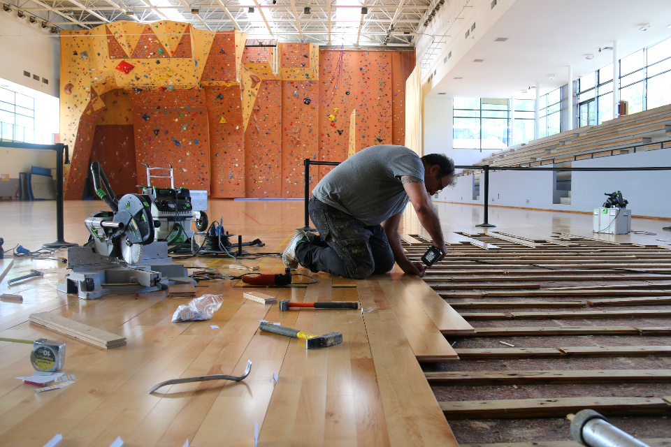 Les réparations du parquet du gymnase Lionel Terray Photo des réparations du parquet du gymnase Lionel Terray. La photo est prise dans le gymnase, nous voyons le mur d'escalade en fond. Au premier plan nous voyons un technicien en train de changer les lames du sol.