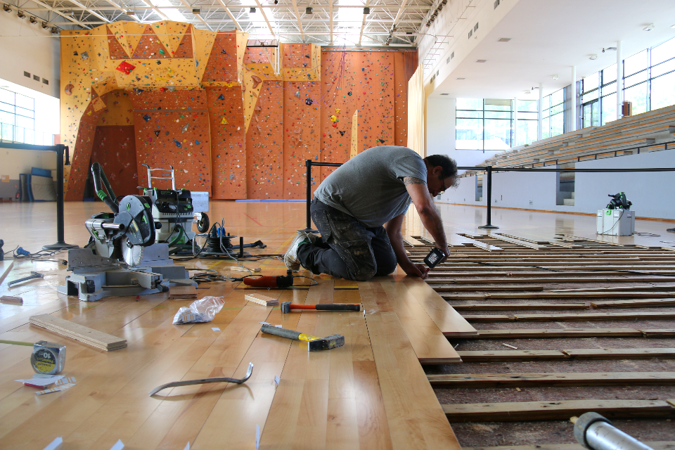 Photo des réparations du parquet du gymnase Lionel Terray. La photo est prise dans le gymnase, nous voyons le mur d'escalade en fond. Au premier plan nous voyons un technicien en train de changer les lames du sol.