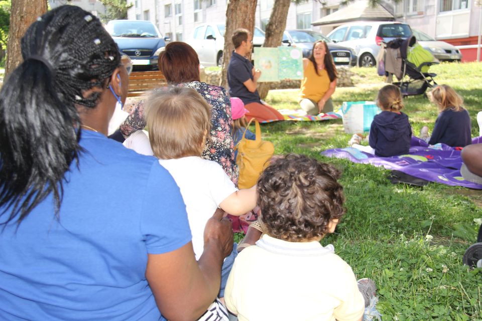 Petite enfance : Les RAM hors les murs Photo prise lors d'un temps de lecture hors les murs proposé par les relais assistantes maternelles (RAM). Nous voyons des parents et des enfants de dos. Face à eux des animateurs des RAM leur lisent une histoire.
