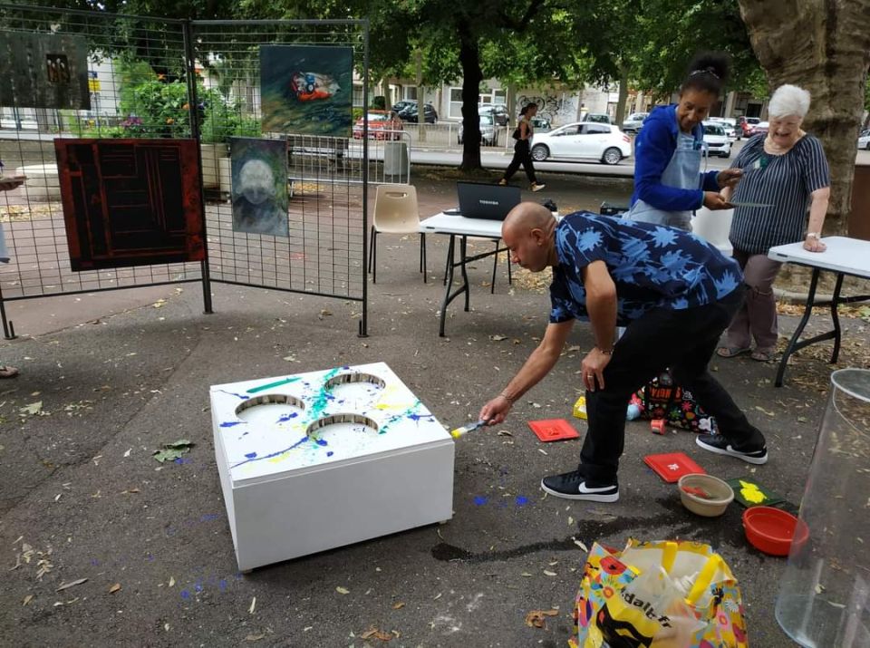 Reg'Arts a renoué le lien Photo prise lors d'un atelier artistique. Un visiteur participe à la création d'un totem à base de matériaux de récupérations. On le voit peindre sur un meuble. D'autres habitant-es l'observent.