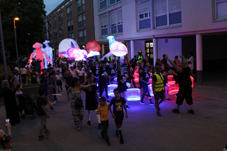 Photo prise à l'occasion de l'événement Carna’light.  Nous voyons des ballons géants lumineux en forme de boule, de baleine. des mascottes géantes en carton. Les participants sont habillés de vêtements blanc avec des marques fluorescentes dessus. Ils se reflètent dans la nuit.  Le public nombreux les entourent pour observer le spectacle. Au centre, la batukavi et ses percussions. Les tambours sont lumineux, aux couleurs blanche et rouge. La batukavi s'élance dans sa déambulation.