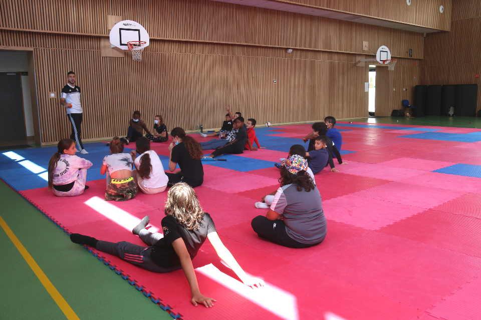 Photo prise avant une activité sportive. Des jeunes garçons et jeunes filles sont assis sur des tapis et un éducateur sportif se tient debout face à eux et leur explique les consignes.