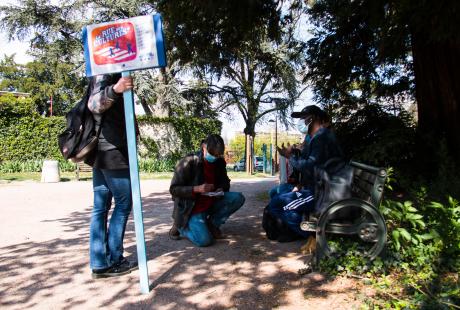 Marie Mazille est assise sur un banc avec un accordéon et avec un habitant.  Fabrice Vigne est agenouillé devant eux et écrit une chanson 