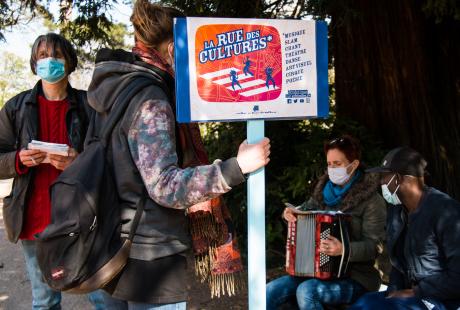 Marie Mazille est assise sur un banc avec un accordéon et avec un habitant.  Fabrice Vigne est debout devant eux. Une accompagnatrice tient le panneau "La Rue des Cultures"