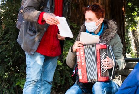 Marie Mazille est assise sur un banc avec un accordéon.  Fabrice Vigne est debout à côté d'elle. Ils échangent sur leur création pour les mutualiser. 