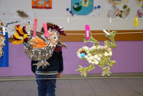 Photo de la salle d’Estienne d’Orves. Des fils sont étendus entre les poteaux et nous voyons des dessins d'enfants exposés, accrochés aux fils avec des pinces. Gros plan sur des dessins d'enfants avec un enfant en arrière plan.