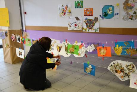 Photo de la salle d’Estienne d’Orves. Des fils sont étendus entre les poteaux et nous voyons des dessins d'enfants exposés, accrochés aux fils avec des pinces. Une agente est en train de tous les accrocher.