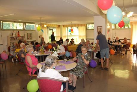 Photo prise à la fête de l'été de la Résidence autonomie Maurice-Thorez. Nous voyons les résidant-es seniors assis autour de tables rondes. des ballons colorés sont suspendus au plafond