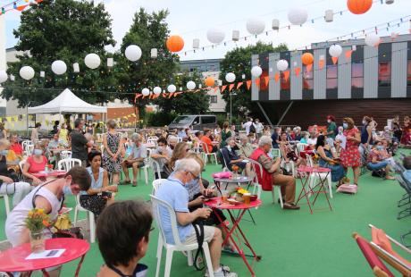 Photo de la présentation de saison la Rampe-La Ponatière pour la saison 2021-2022. Celle-ci se déroulait en extérieur. Le parking a été recouvert d'un tapis vert, effet herbe. Des tables de jardin et des chaises sont installées, des fanions et lampions blanc et orange (aux couleurs de la saison) sont suspendus en hauteur. Nous voyons les habitant-es s'installer sur les tables.