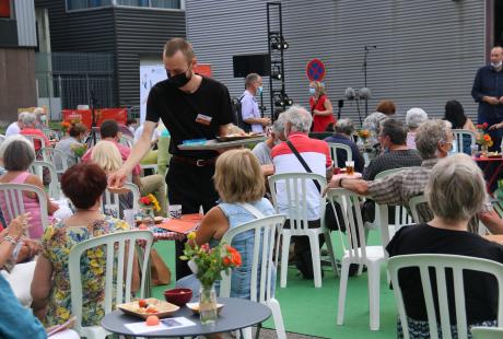 Photo de la présentation de saison la Rampe-La Ponatière pour la saison 2021-2022. Celle-ci se déroulait en extérieur. Le parking a été recouvert d'un tapis vert, effet herbe. Des tables de jardin et des chaises sont installées, des fanions et lampions blanc et orange (aux couleurs de la saison) sont suspendus en hauteur. Nous voyons le public installé sur les tables.