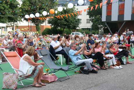 Photo de la présentation de saison la Rampe-La Ponatière pour la saison 2021-2022. Celle-ci se déroulait en extérieur. Le parking a été recouvert d'un tapis vert, effet herbe. Des tables de jardin et des chaises sont installées, des fanions et lampions blanc et orange (aux couleurs de la saison) sont suspendus en hauteur. Nous voyons les habitant-es installés sur les tables. Au milieu de la foule nous apercevons le maire et quelques élu-es.