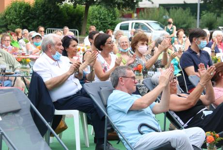   Photo de la présentation de saison la Rampe-La Ponatière pour la saison 2021-2022. Celle-ci se déroulait en extérieur. Le parking a été recouvert d'un tapis vert, effet herbe. Des tables de jardin et des chaises sont installées, des fanions et lampions blanc et orange (aux couleurs de la saison) sont suspendus en hauteur. Nous voyons le maire, Renzo Sulli, la première adjointe, Amandine Demore et l'adjointe à la Culture, Jacqueline Madrennes installés dans le public.