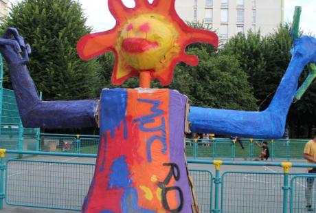 Photo prise à l'occasion de l'événement Carna’light. Photo d'une mascotte géante en carton. Elle a une tête de soleil et les bras levés vers le haut. 