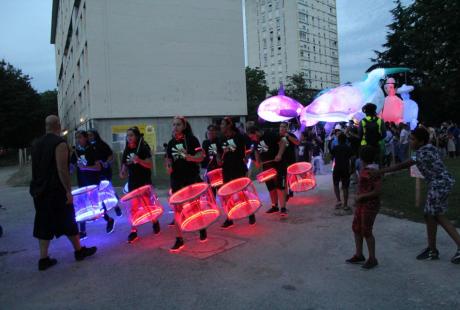 Photo prise à l'occasion de l'événement Carna’light.  Nous voyons des ballons géants lumineux en forme de boule, de baleine. des mascottes géantes en carton. Les participants sont habillés de vêtements blanc avec des marques fluorescentes dessus. Ils se reflètent dans la nuit.  Le public nombreux les entourent pour observer le spectacle. Au centre, la batukavi et ses percussions. Les tambours sont lumineux, aux couleurs blanche et rouge.