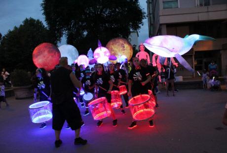 Photo prise à l'occasion de l'événement Carna’light.  Nous voyons des ballons géants lumineux en forme de boule, de baleine. des mascottes géantes en carton. Les participants sont habillés de vêtements blanc avec des marques fluorescentes dessus. Ils se reflètent dans la nuit.  Le public nombreux les entourent pour observer le spectacle. Au centre, la batukavi et ses percussions. Les tambours sont lumineux, aux couleurs blanche et rouge.