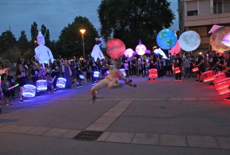 Photo prise à l'occasion de l'événement Carna’light.  Nous voyons des ballons géants lumineux en forme de boule, de baleine. des mascottes géantes en carton. Les participants sont habillés de vêtements blanc avec des marques fluorescentes dessus. Ils se reflètent dans la nuit.  Le public nombreux les entourent pour observer le spectacle. Au centre, un artiste fait une démonstration de hip hop