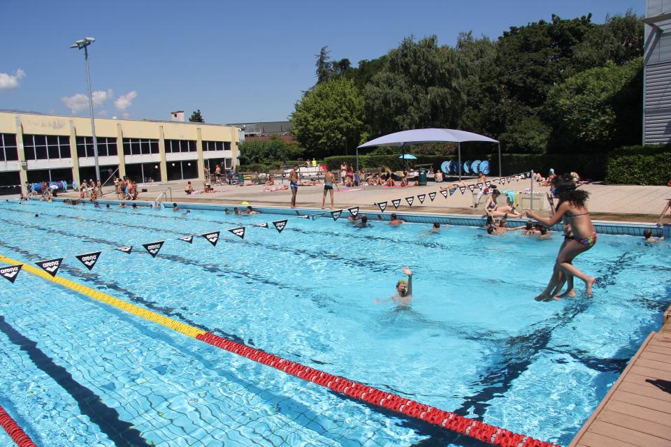 Destination été ! Vue sur le bassin extérieur du stade nautique. Deux enfants sautent du ponton dans la piscine.