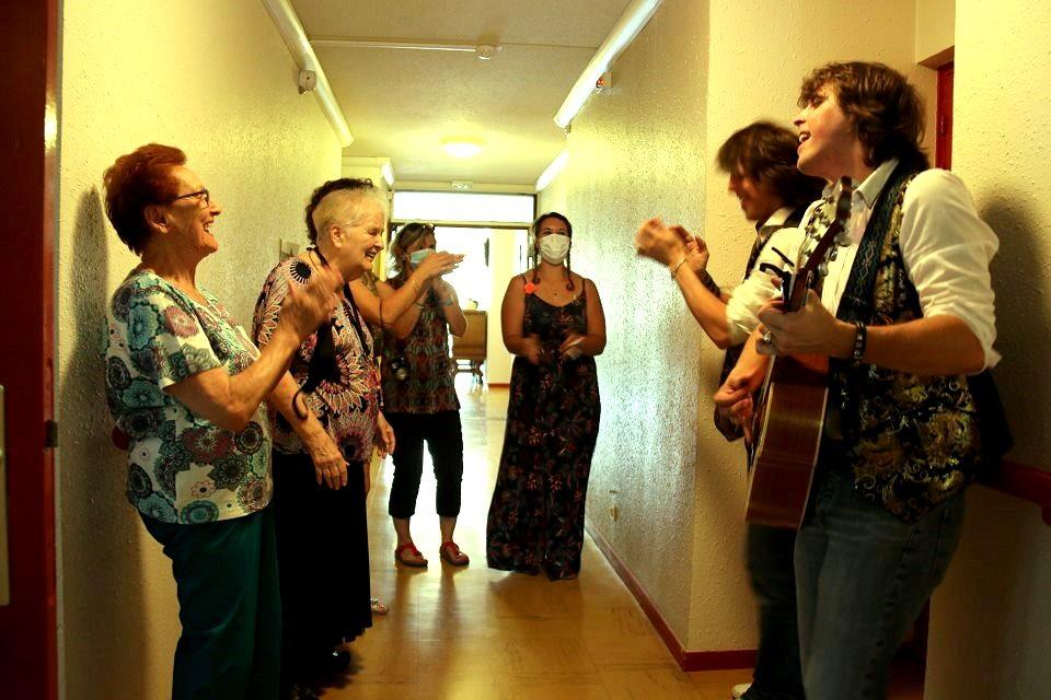 Un guitariste et un chanteur jouent dans le couloir de la Résidence Maurice-Thorez. Plusieurs personnes âgées chantent, sourient et applaudissent.