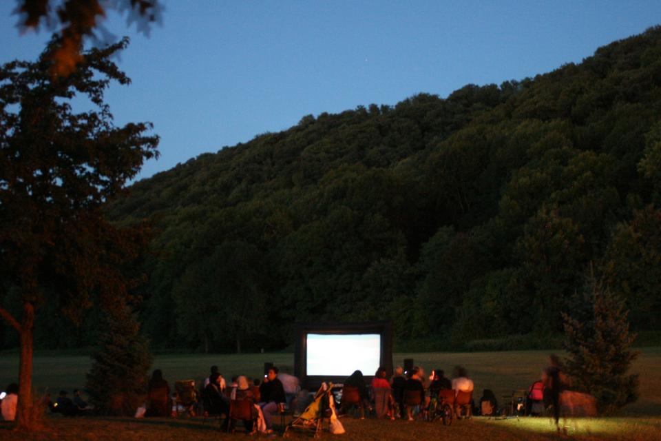 Une grande toile blanche est installée au milieu du parc. Le public s'installe en vue de la projection nocturne.