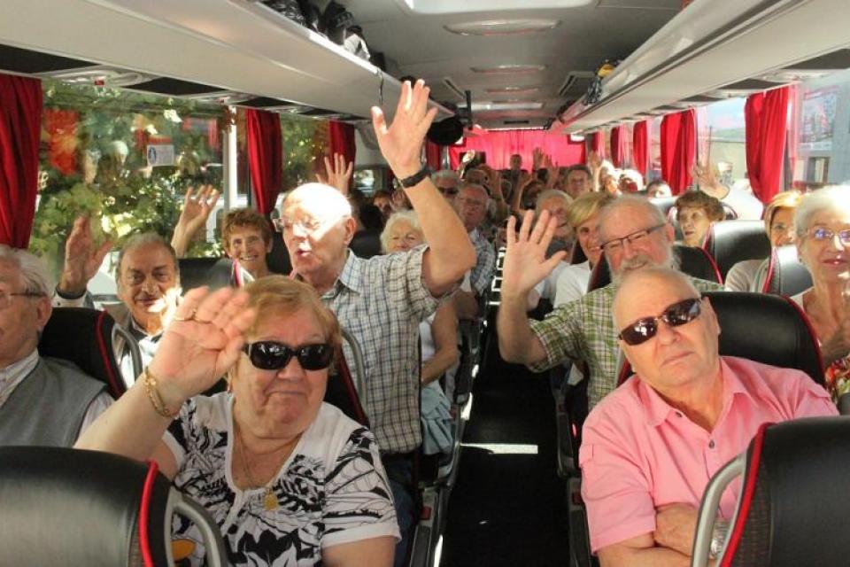 Plusieurs retraité-es sont asis dans un bus. Ils font signe au photographe et sourient avant leur départ en vacances.