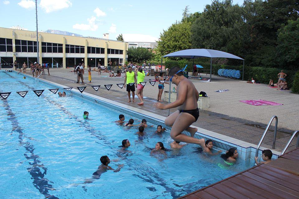 Vue sur le bassin extérieur du stade nautique. Un groupe d'adolescent-es est dans l'eau, accroché au bord du bassin. Un garçon saute du bord de la piscine pour plonger à côté de ses copains.