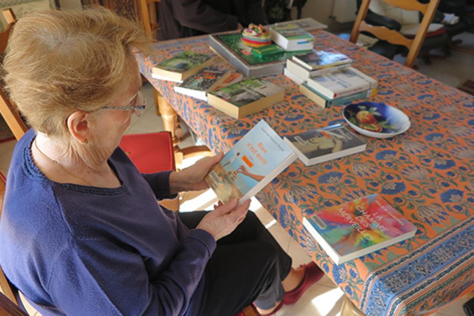 Une personne âgée est assise à une table. Elle regarde la couverture d'un livre. Plusieurs livres sont posés sur la table devant elle.