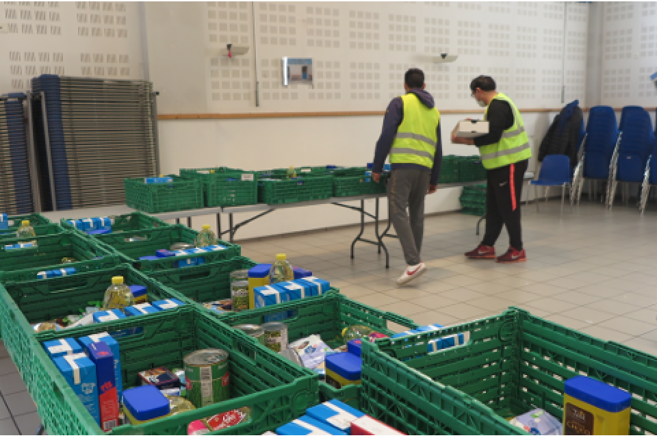 Les bénévoles organisent la distribution de colis alimentaire. Plusieurs cagettes de produits alimentaires sont installées sur des tables.