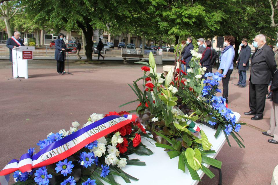 76e anniversaire du 8-mai-1945 Au premier plan nous voyons les gerbes de fleurs qui seront déposées sur le monument aux morts. En arrière plan nous voyons les représentant-es d'associations et les élu-es, aligné-es, en train d"écouter le discours du Maire, qui se tient derrière un pupitre.