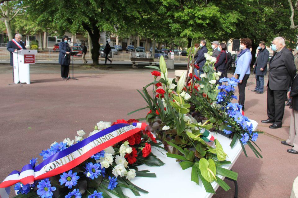 Au premier plan nous voyons les gerbes de fleurs qui seront déposées sur le monument aux morts. En arrière plan nous voyons les représentant-es d'associations et les élu-es, aligné-es, en train d"écouter le discours du Maire, qui se tient derrière un pupitre.