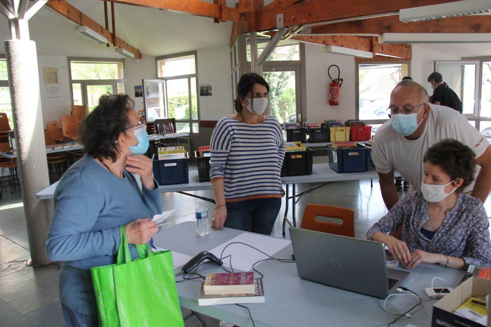 Photo de la bibliothèque éphémère. Nous voyons plusieurs agents derrière un ordinateur qui discutent avec une habitante. En arrière plan nous voyons des caisses remplies de livres, disposées sur des tables.