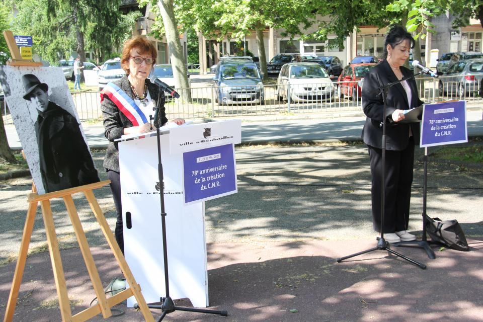 78e anniversaire de la création du CNR. Photo prise place de la Libération. Nous voyons sur un grand présentoir, une photo en noir et blanc de Jean Moulin. A côté de celui-ci, Jacqueline Madrennes, Adjointe au travail de mémoire, s'exprime derrière un pupitre avec un micro.