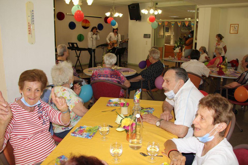 Photo prise à la fête de l'été de la Résidence autonomie Maurice-Thorez. Nous voyons les résidant-es seniors assis autour de tables rondes, ils regardent une guitariste et une chanteuse qui donnent un spectacle.